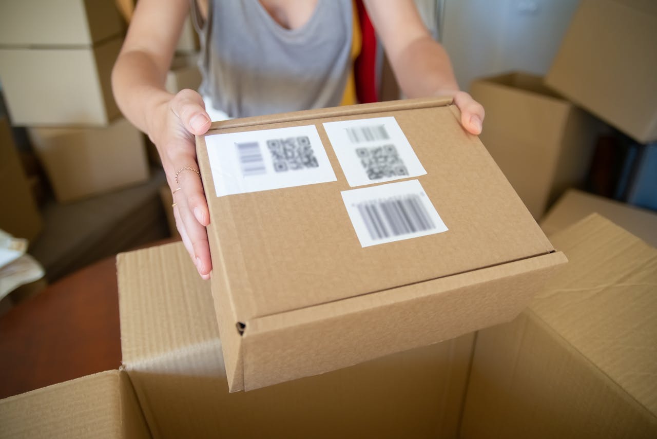 Home Close-up of a person holding a cardboard box with barcode labels, indoors setting.